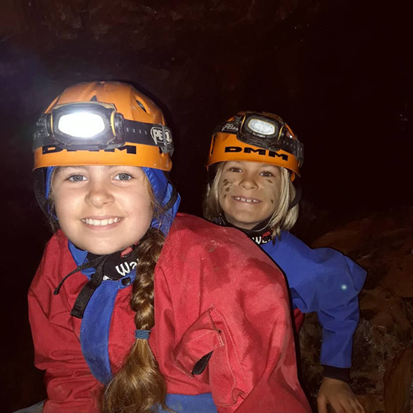 Young siblings smiling at the camera. They have helmets and lights on as they're in a dark cave.